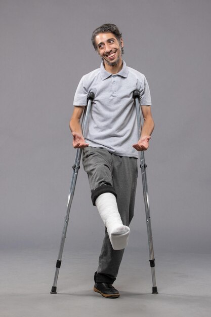 Front view young man using crutches due to broken foot on grey wall leg accident disabled broke damage broken