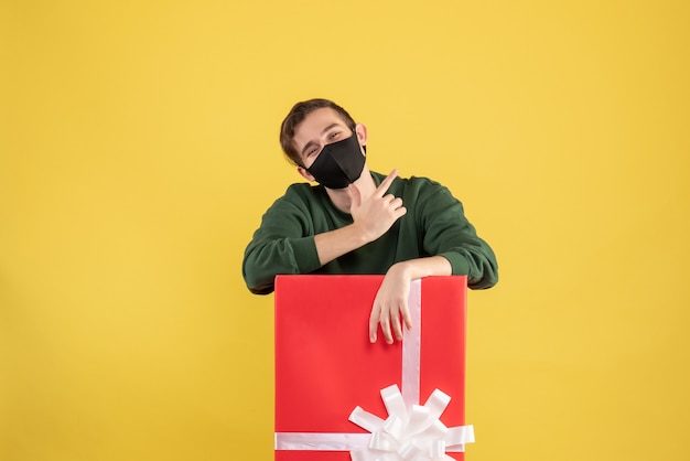 Front view young man with black mask standing behind big giftbox on yellow 