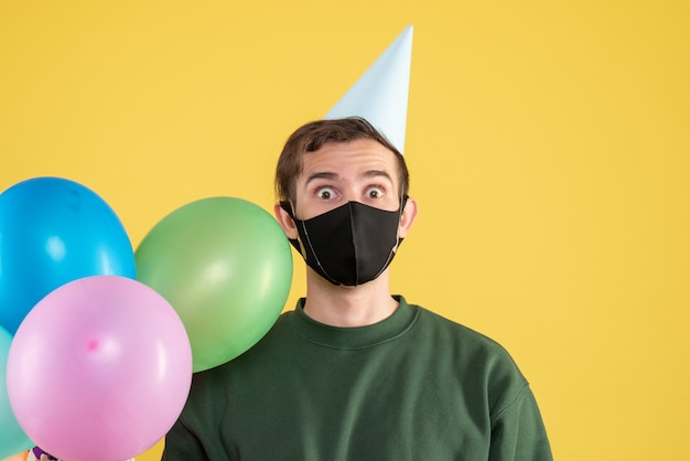 Front view young man with party cap and black mask colorful balloons on yellow 