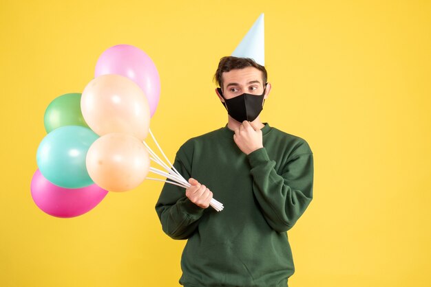 Front view young man with party cap and colorful balloons standing on yellow 