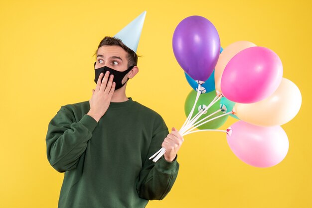 Front view young man with party cap and colorful balloons standing on yellow 