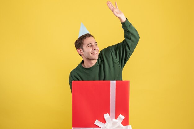 Front view young man with party cap hailing someone standing behind big giftbox on yellow background