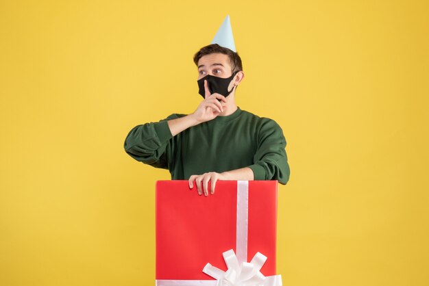 Front view young man with party cap making shh sign standing behind big giftbox on yellow background