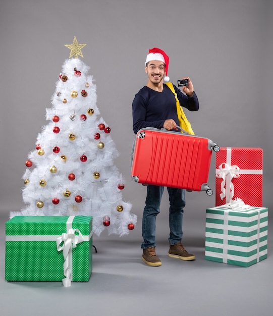 Free Photo front view young man with santa hat holding red suitcase showing a card around presents on grey isolated