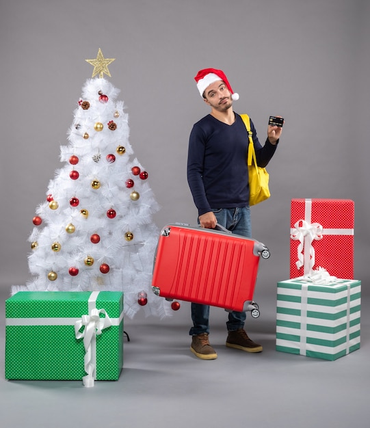 Free photo front view young man with santa hat holding red suitcase showing a card around presents on isolated