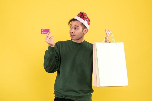 Free photo front view young man with santa hat holding shopping bags and card standing on yellow background copy place