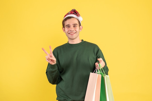 Front view young man with santa hat holding shopping bags making victory sign on yellow background