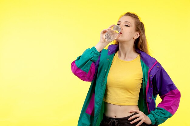 A front view young modern woman in yellow shirt black trousers and colorful jacket drinking water