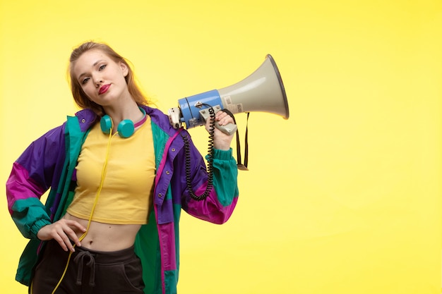 Free photo a front view young modern woman in yellow shirt black trousers and colorful jacket with colored earphones holding megaphone posing