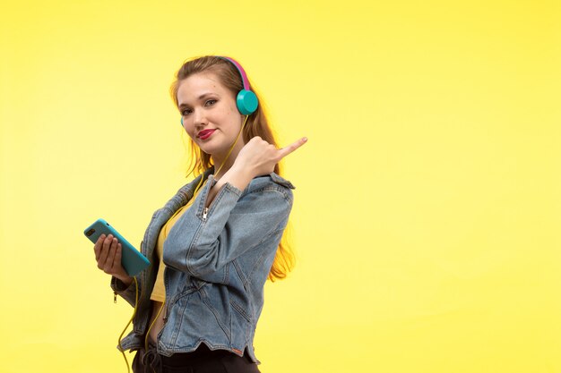 A front view young modern woman in yellow shirt black trousers and jean coat with colored earphones using phone posing