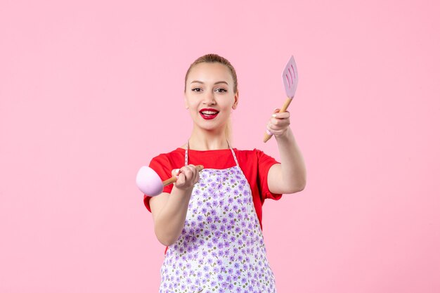 Front view young pretty housewife in cape with spoons on pink wall