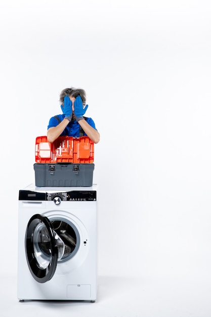 Front view of young repairman covering his face with hands washing machine on white wall