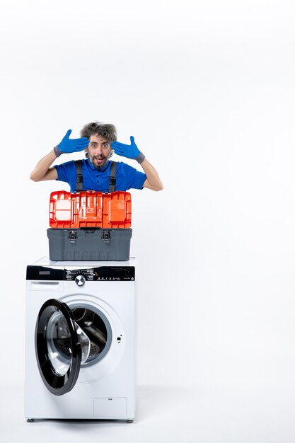 Front view of young repairman putting hands to his temple behind washing machine on white wall