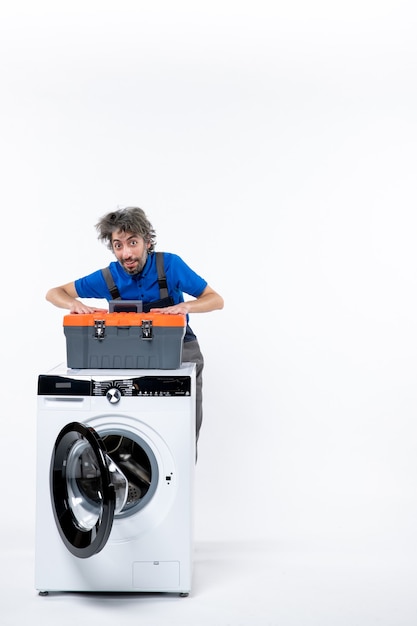 Front view of young repairman putting hands on his tools bag standing behind washing machine on white wall