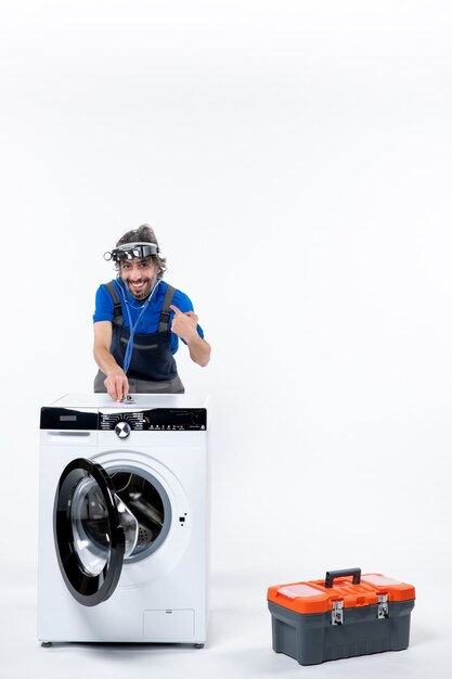 Front view of young repairman with head lamp putting stethoscope on washer pointing at himself on white wall