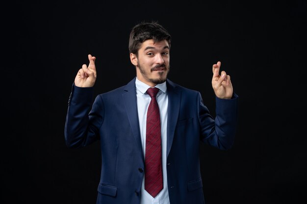 Front view of young and satisfied bearded man crossing his fingers on isolated dark wall