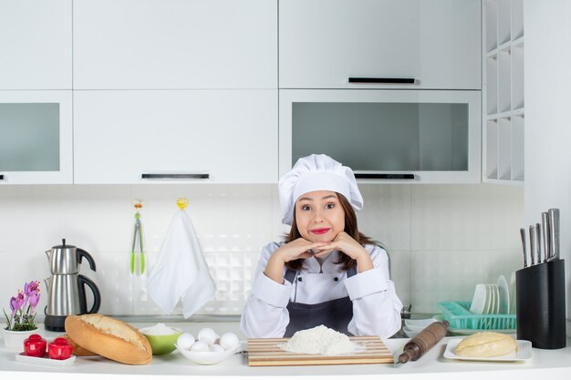 Front view of young smiling female chef in uniform standing behind the table with cutting board foods putting hands under chin in the white kitchen