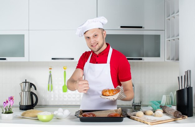 Front view of young smiling male chef wearing holder holding one of freshly-baked pastries and making ok gesture in the white kitchen