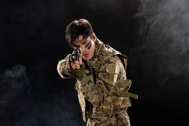 Front view of young soldier in camouflage aiming gun on black wall