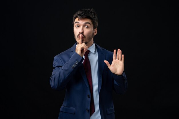 Front view of young surprised bearded man making silence gesture and showing five on isolated dark wall