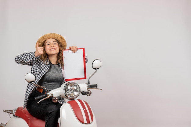 Front view young traveller girl on moped holding red clipboard giving thumbs up