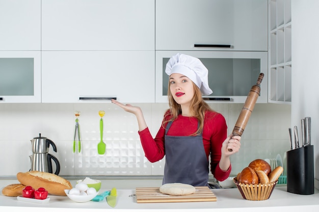 Front view young woman in cook hat and apron holding rolling pin in the kitchen