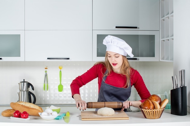 Front view young woman in cook hat and apron rolling dough in the kitchen