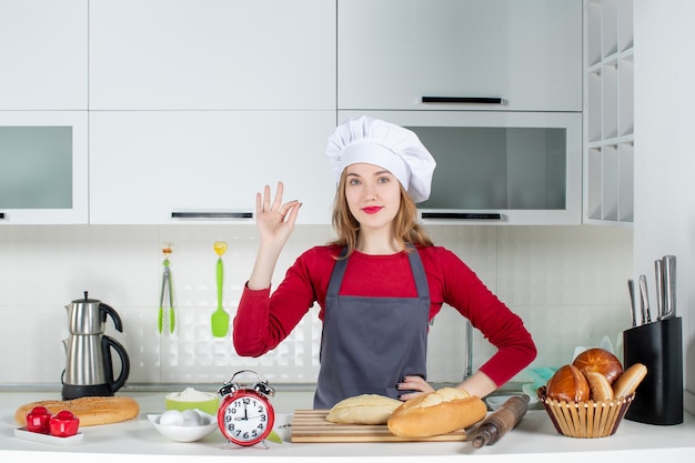 Front view young woman in cook hat and apron standing behind table in the kitchen