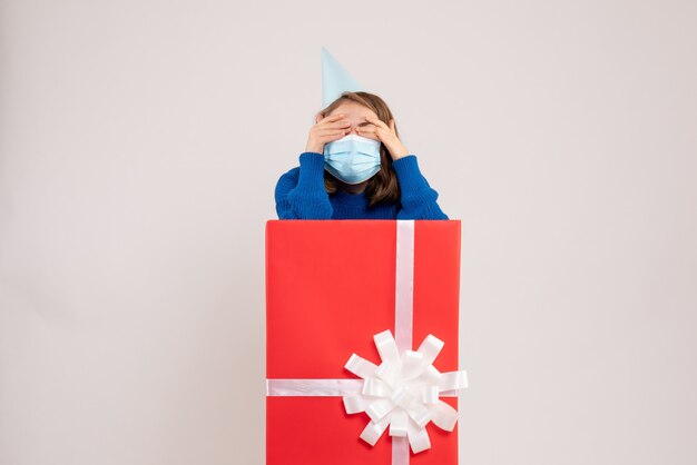 Front view of young woman inside present box in mask on white wall