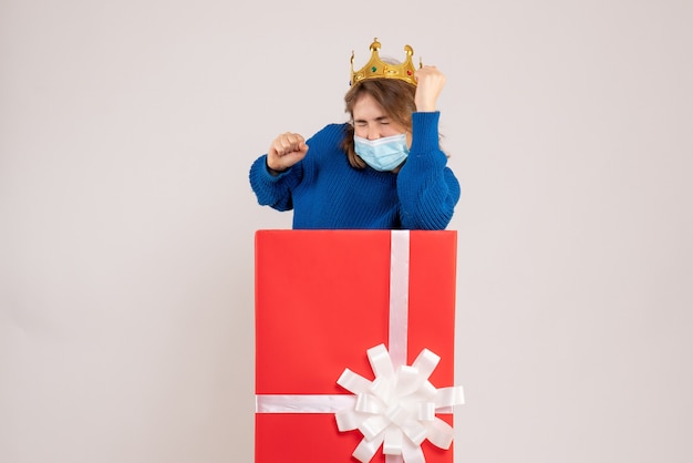 Front view of young woman inside present box in sterile mask on white wall