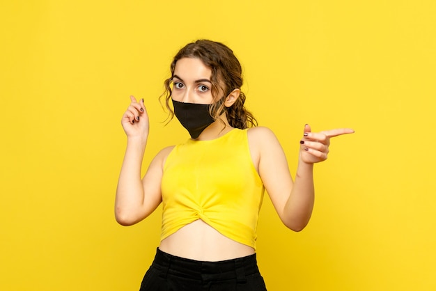 Front view of young woman pointing in black mask on yellow wall