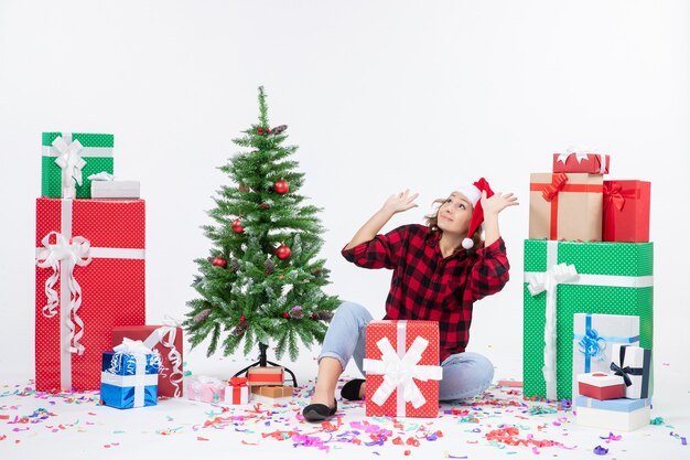 Front view of young woman sitting around christmas presents and little holiday tree on white wall