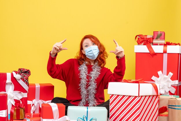 Front view of young woman sitting around christmas presents in mask on yellow wall