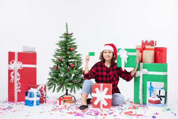 Front view of young woman sitting around presents holding green bank card on white wall