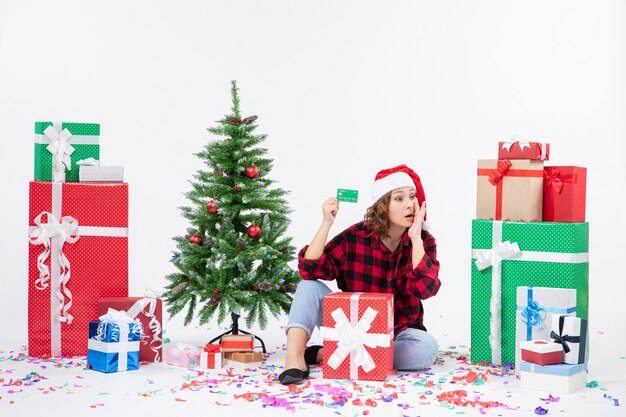 Front view of young woman sitting around presents holding green bank card on white wall