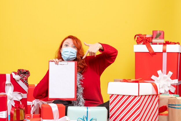 Front view of young woman sitting around presents in mask with file note on a yellow wall