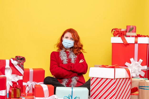 Front view of young woman sitting around presents in mask on a yellow wall