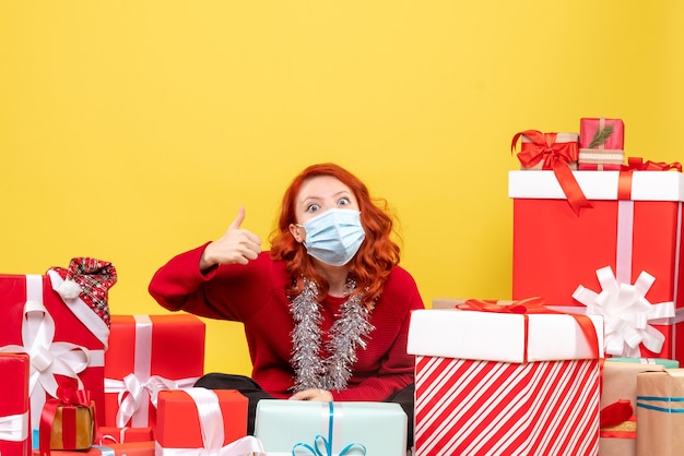 Front view of young woman sitting around presents in mask on yellow wall