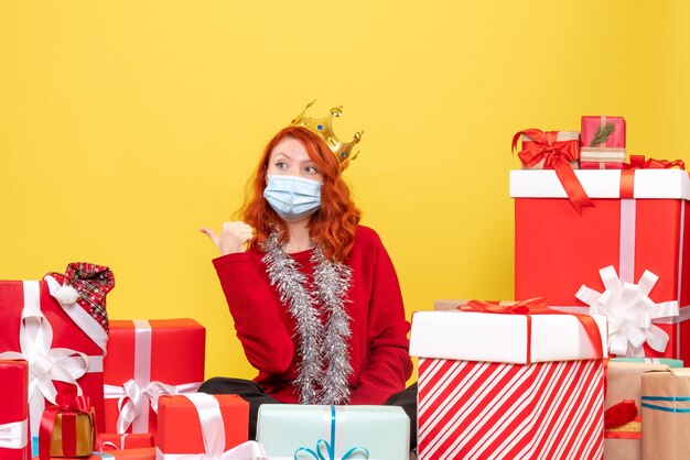 Front view of young woman sitting around presents in sterile mask on the yellow wall