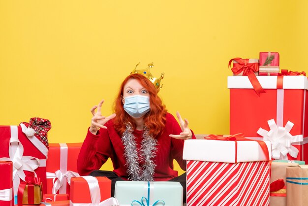 Front view of young woman sitting around presents in sterile mask on yellow wall