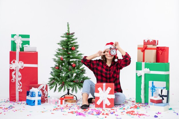 Front view of young woman sitting around xmas presents holding clocks on white wall