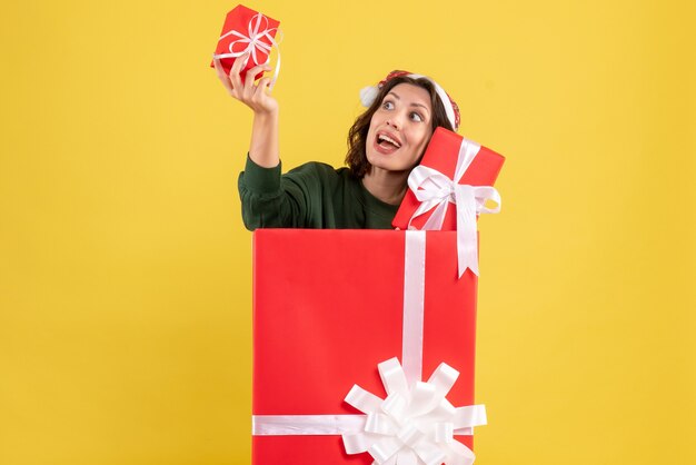 Front view of young woman standing inside box with xmas presents on yellow wall