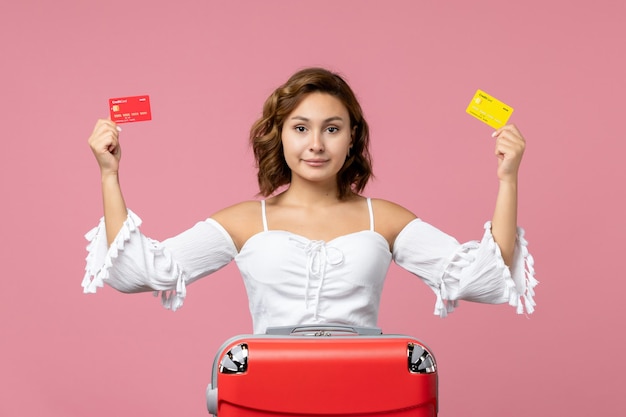 Front view of young woman with red vacation bag holding bank cards on pink wall