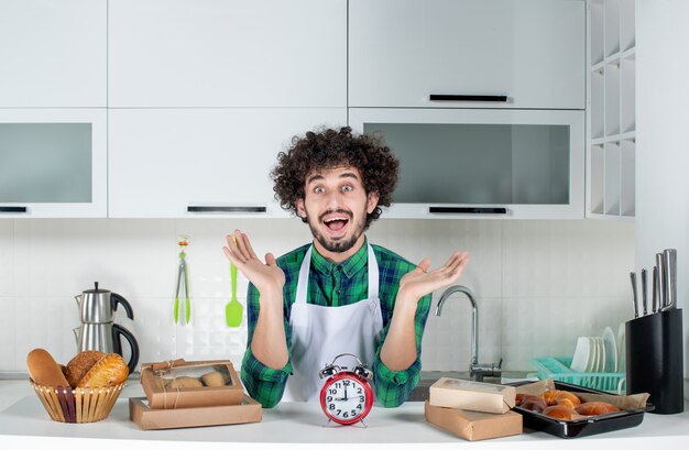 Free photo front view of young wondering man standing behind the table clock various pastries on it in the white kitchen