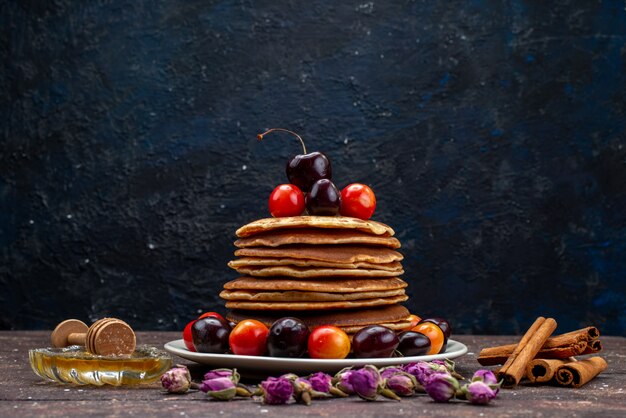 A front view yummy pancakes with cherries inside white plate with flowers and cinnamon on the dark desk fruit pancake