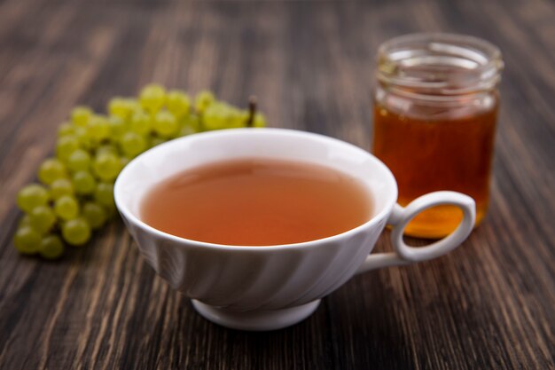 Frontal view cup of tea with green grapes and honey in a jar on wooden background