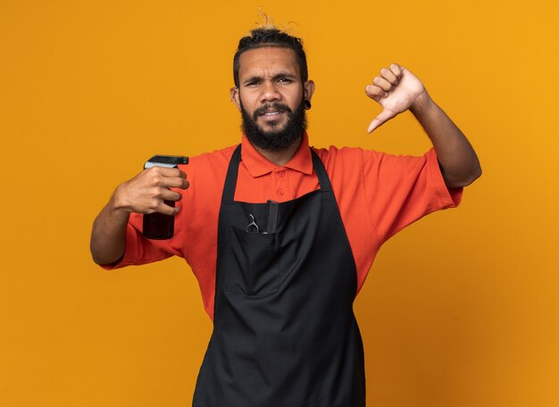 Frowning young afro-american male barber wearing uniform holding hair spray  showing thumb down isolated on orange wall