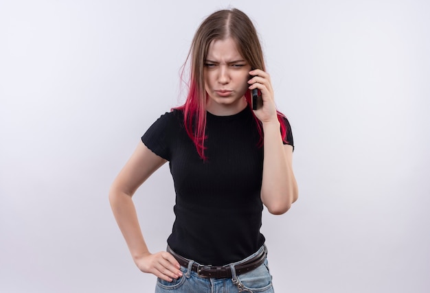 Frowning young beautiful woman wearing black t-shirt speakes on phone putting her hand on hip on isolated white wall