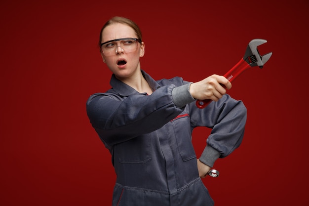 Free Photo frowning young blonde female engineer wearing uniform and safety glasses holding wrenches keeping hand on waist looking and pointing at side with wrench 