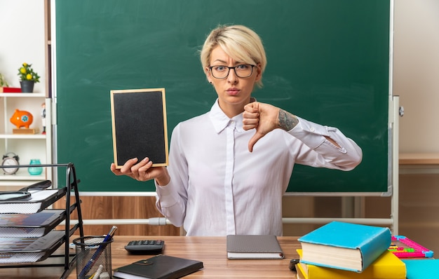 Free photo frowning young blonde female teacher wearing glasses sitting at desk with school tools in classroom showing mini blackboard and thumb down